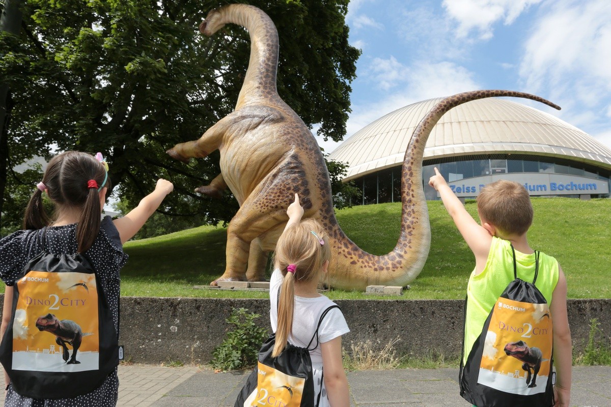 Direkt nach dem Aufbau des Apatosaurus vor dem Planetarium haben sich die Kinder Sophia, Elias und Evelina (v.li.) den Dinosaurier angeschaut. Mit dabei hatten sie den Dino City-Rucksack, der in der Bochum Touristinfo erhältlich ist.