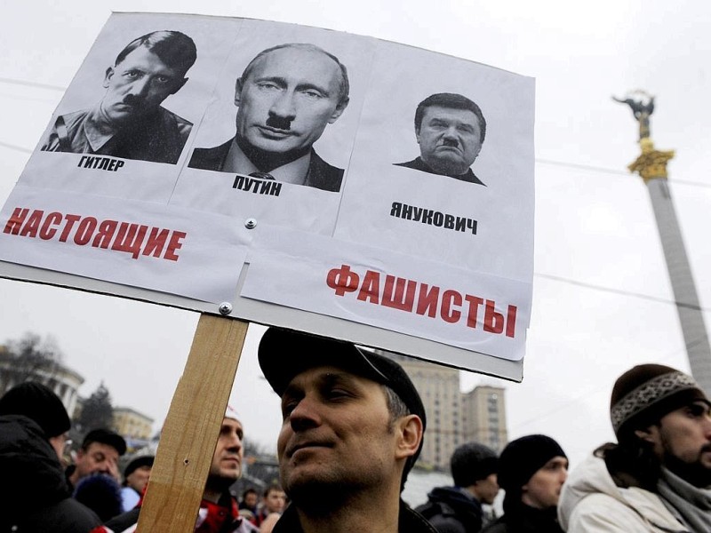 Während der Demonstration hält ein Mann ein Schild hoch, auf dem Wladimir Putin und Viktor Janukowitsch neben Adolf Hitler zu sehen sind.