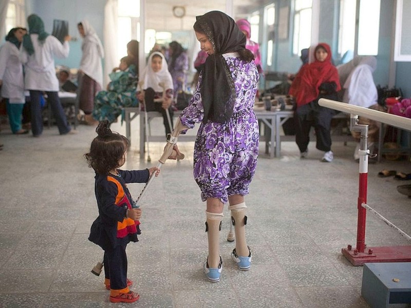 Ein afghanisches Mädchen probiert, auf seinen neuen Bein-Prothesen zu laufen; die kleine Schwester greift nach den Krücken.
