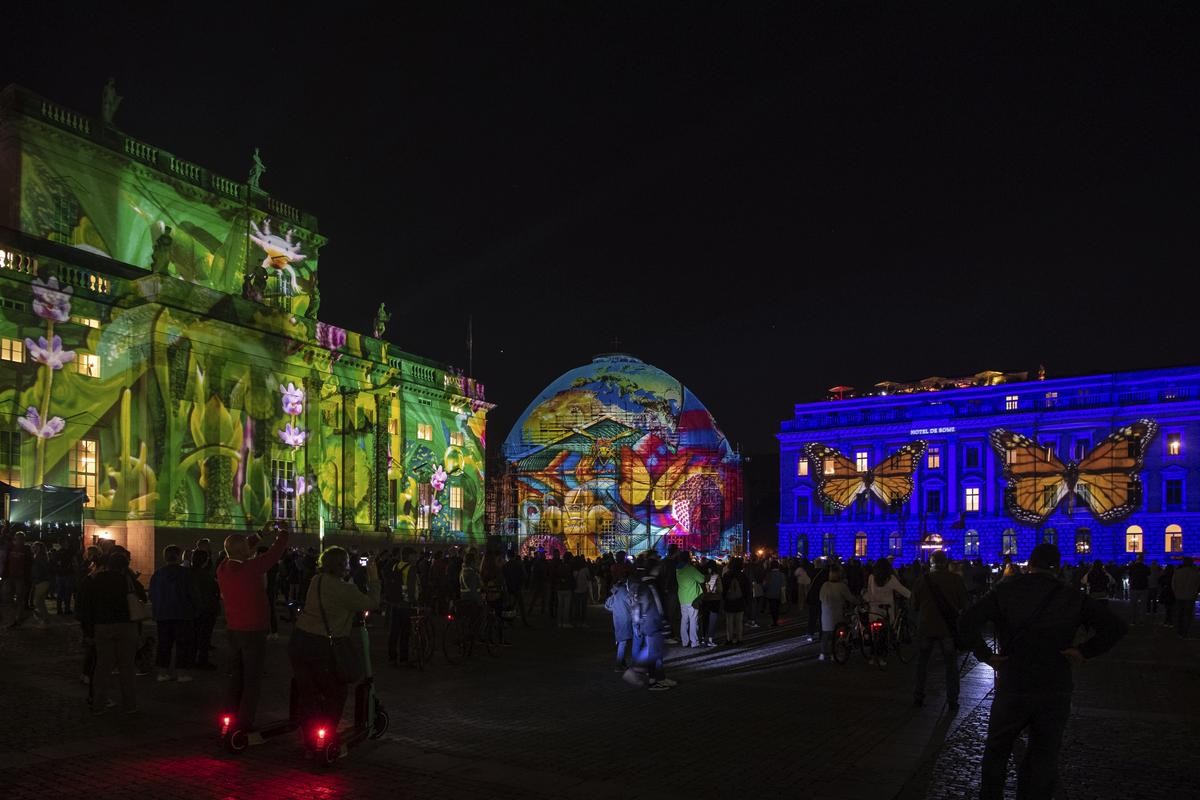 Zahlreiche Menschen stehen zum Auftakt des 17. Festival of Lights 2021 auf dem Bebelplatz. Dort sind alle Gebäude illuminiert.