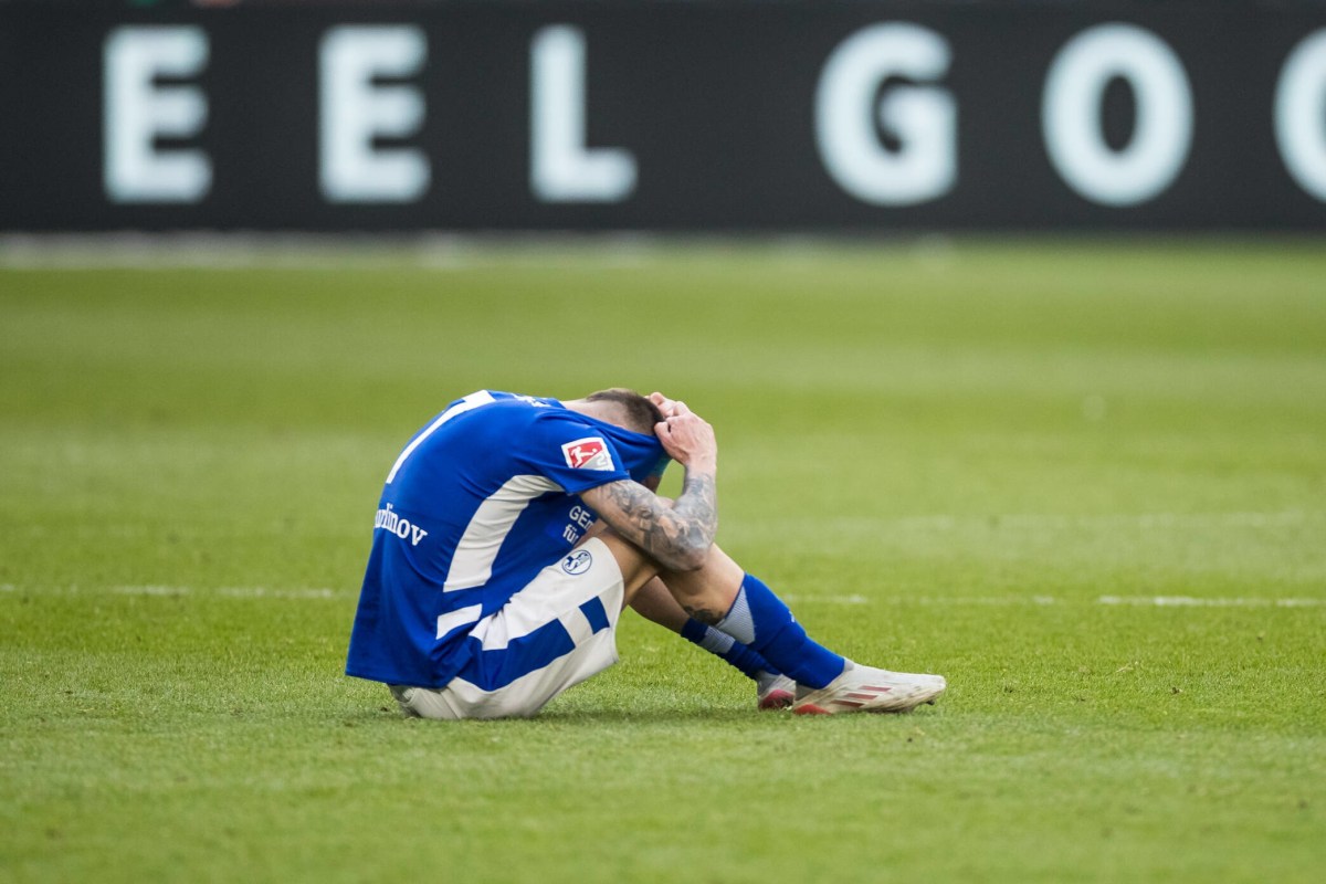 Darko Churlinov sitzt frustriert auf dem Rasen und zieht sich das Trikot des FC Schalke 04 über den Kopf.
