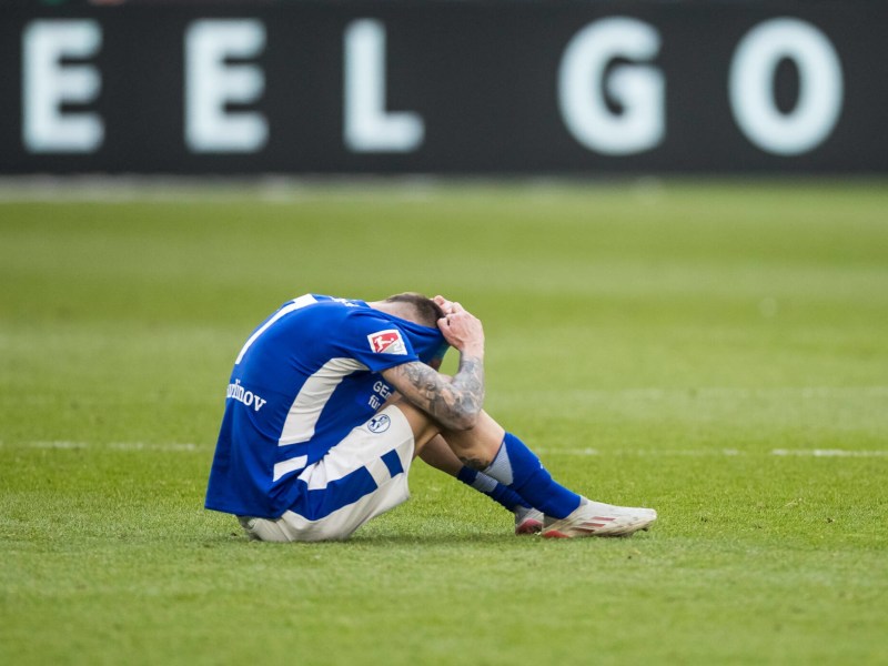 Darko Churlinov sitzt frustriert auf dem Rasen und zieht sich das Trikot des FC Schalke 04 über den Kopf.