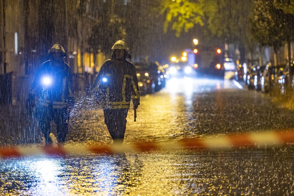 Zwei Feuerwehrmänner laufen in Duisburg über eine überschwemmte Straße mit Flatterband