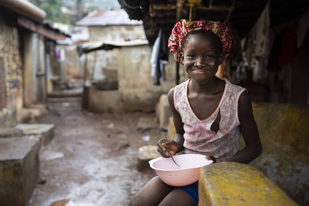 Ein Mädchen mit einem Teller sitzt auf einer Bank einer Armensiedlung in Freetown, Sierra Leone.