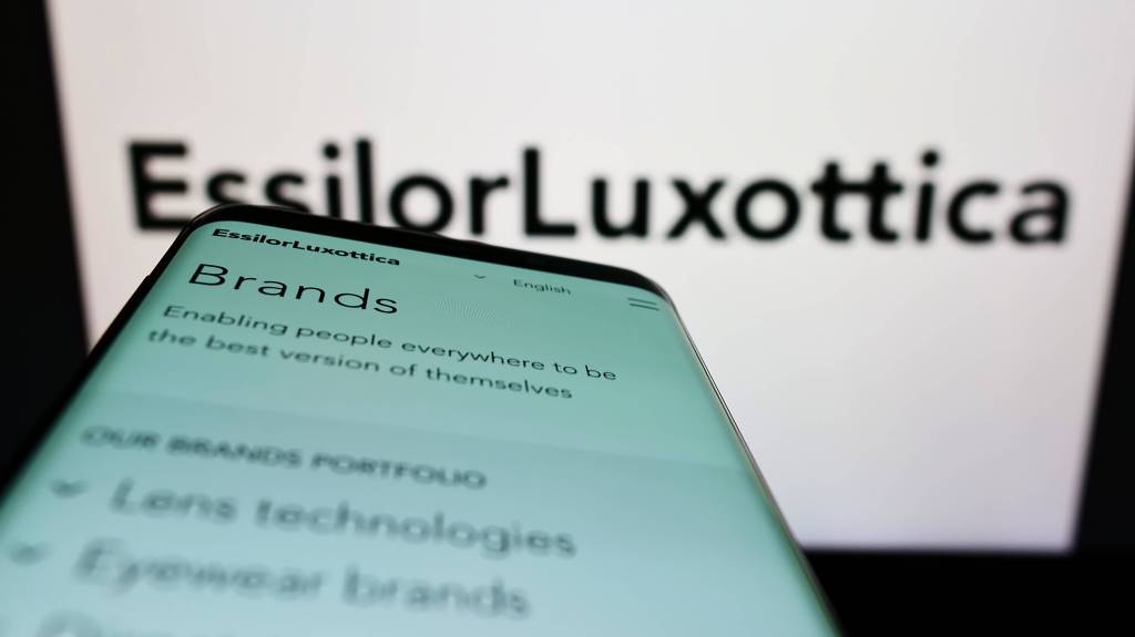 Ein Smartphone wird vor einen Computer-Monitor gehalten, auf dem die Unternehmens-Website von EssilorLuxottica zu sehen ist.