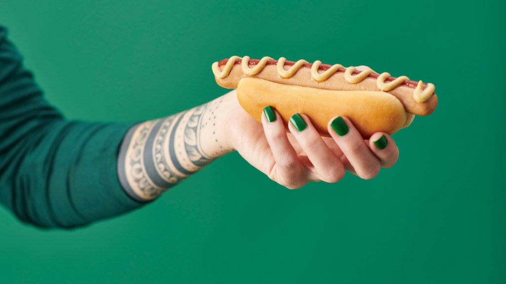 Frau hält Ikea-Hotdog in der Hand vor grünem Hintergrund