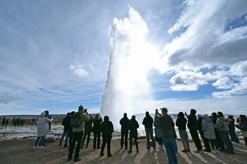 Zahlreiche Menschen stehen unter freiem Himmel vor einem Geysir, der eine Fontäne in die Luft schießt.