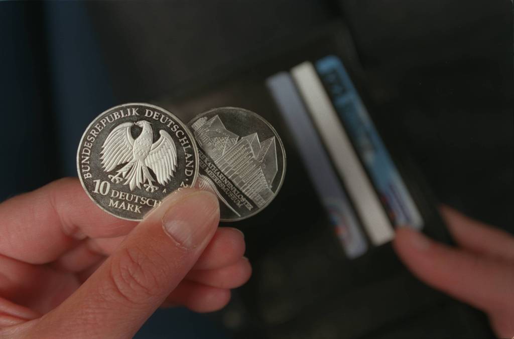 Eine Person hält zwei 1ß-DM-Münzen in der Hand, während im Hintergrund ein Portemonnaie zu sehen ist.
