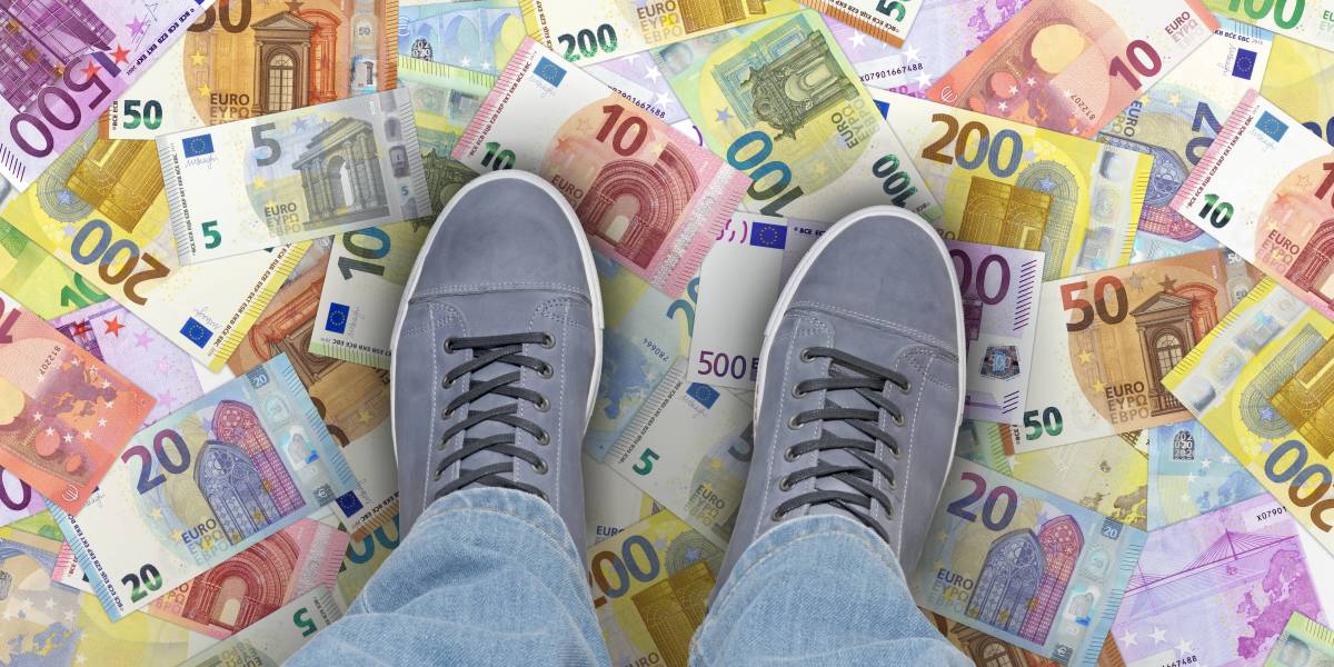 Ein Mensch steht mit beiden Füßen auf Euro-Geldscheinen, die auf dem Boden verteilt liegen.
