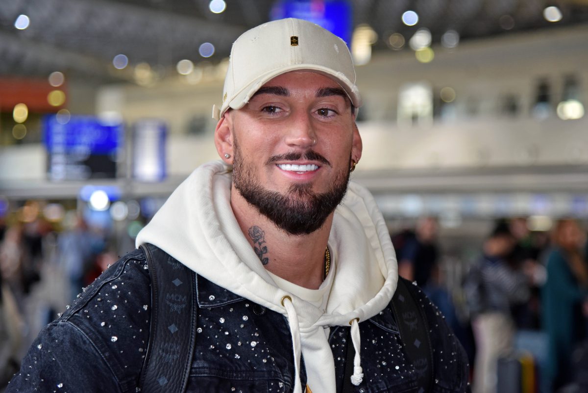 Mike Heiter am Flughafen-terminal. Er trägt eine beige Cap und einen beigen Pullover. Über dem Pullover trägt er eine schwarze Jeansjacke.