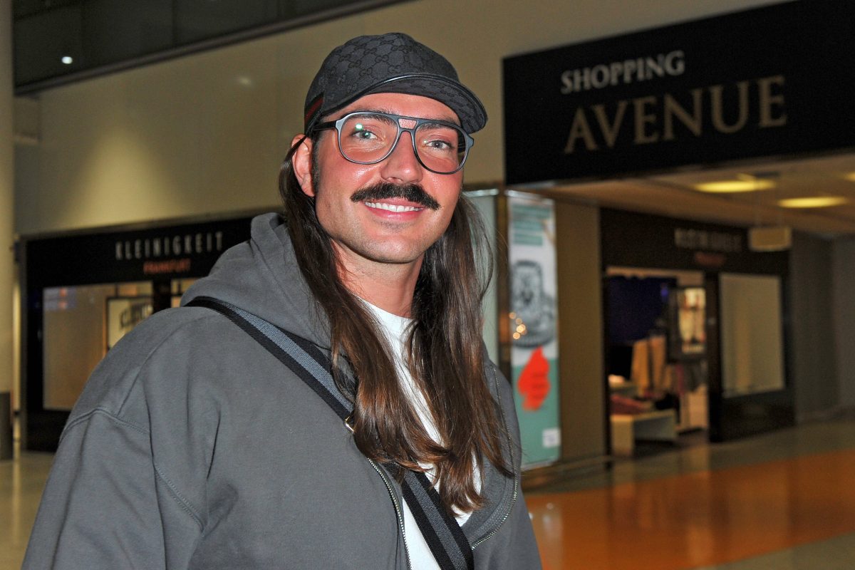 Auf dem Bild ist der Reality-Star Fabio Knez zu sehen. Er steht in der Halle eines Flughafens. Er hat lange braune Haare und trägt eine Mütze. In seinem Gesicht trägt er eine schwarze Retro-Brille und lächelt.