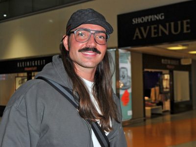 Auf dem Bild ist der Reality-Star Fabio Knez zu sehen. Er steht in der Halle eines Flughafens. Er hat lange braune Haare und trägt eine Mütze. In seinem Gesicht trägt er eine schwarze Retro-Brille und lächelt.