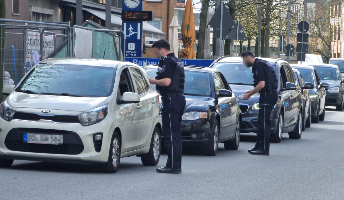 Zwei Polizisten kontrollieren Autos in einer Schlange. Vorne steht ein graues Auto und dahinter mehrere schwarze Autos. Die Polizisten stehen gebeugt vor den offenen Fenstern der ersten beiden Autos in der Schlange.