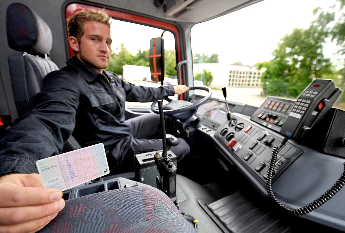 Ein Mann mit blonden Haaren sitzt am Steuer in einem LKW. Er trägt schwarz/graue Arbeitskleidung. Seine linke Hand hat er am Steuer. Mit der rechten Hand zeigt er seinen Führerschein ins Bild.