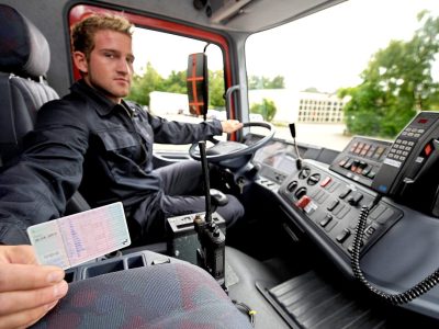 Ein Mann mit blonden Haaren sitzt am Steuer in einem LKW. Er trägt schwarz/graue Arbeitskleidung. Seine linke Hand hat er am Steuer. Mit der rechten Hand zeigt er seinen Führerschein ins Bild.