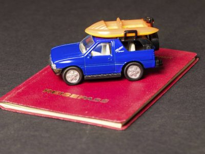 Auf dem Bild sieht man einen Reisepass. Auf dem Reisepass steht ein blaues Spielzeugauto. Auf dem Auto ist außerdem ein Surfbrett.