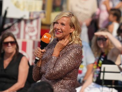 Auf dem Bild zu sehen ist die Moderatorin Andrea Kiewel. In ihrer Hand hat sie ein orangenes Mikrofon mit der Aufschrift "ZDF". Sie träht ein goldenes Kleid mit Glitzer. Zudem lächelt sie und hat schulterlanges blond gelocktes Haar.
