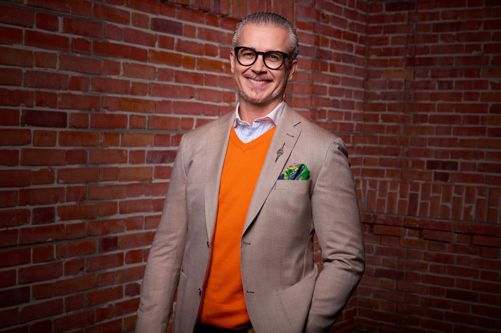 Anaisio Guedes mit orangenem Pullover und beigem Sakko.